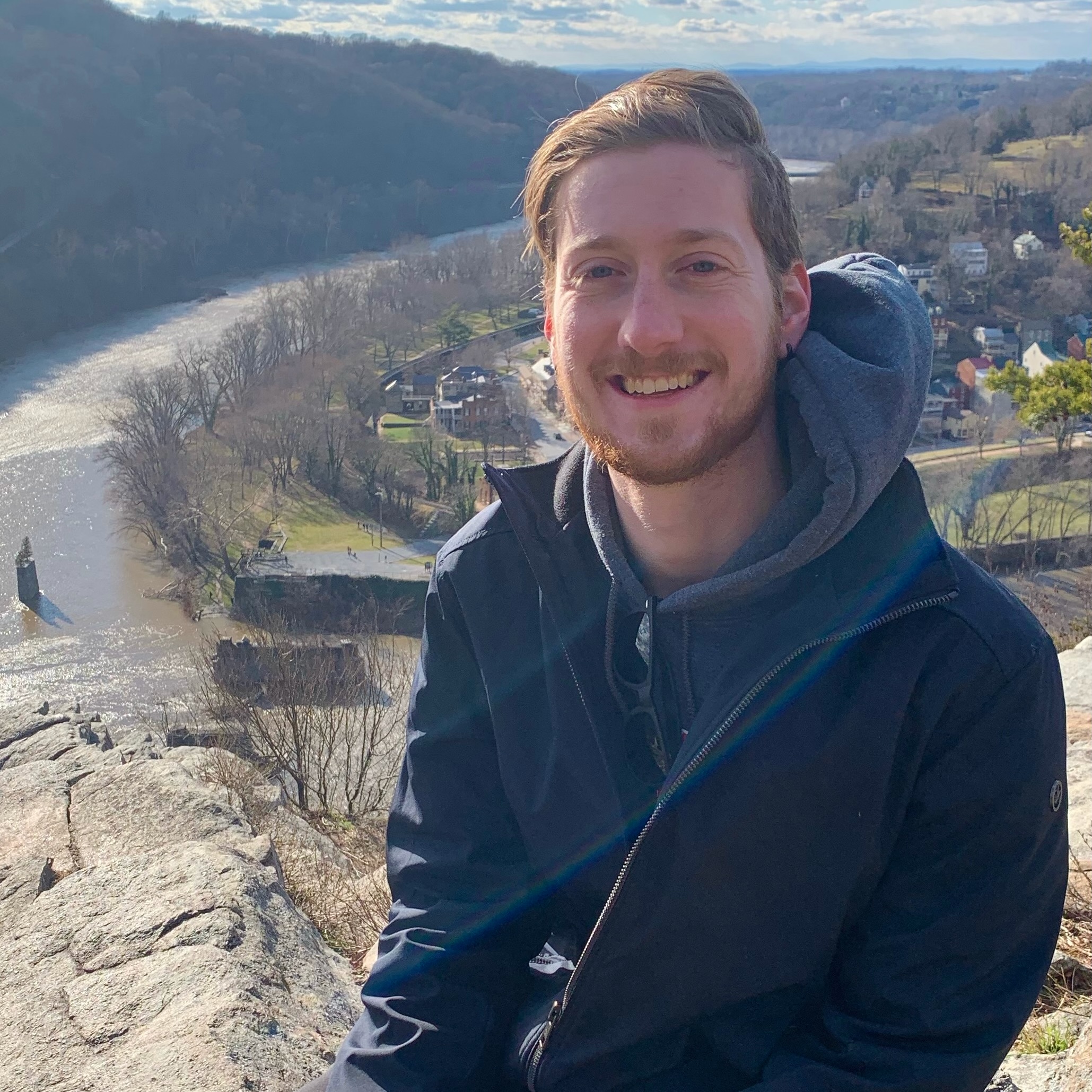 A young man with a short beard, sitting on a mountain above a bend in a river, perhaps Harper's Ferry.